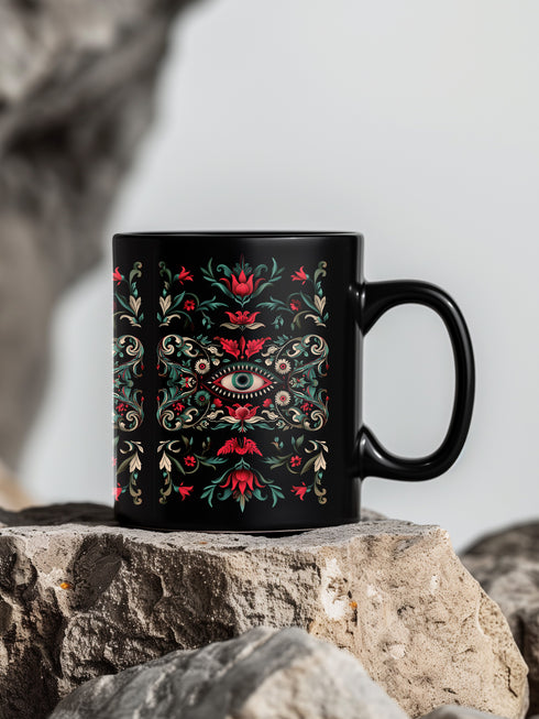 A black ceramic mug featuring a symmetrical folk art design with a central mystic eye motif in turquoise and coral. The design includes ornate scrollwork, red tulip-like flowers, sage green leaves, and cream flourishes arranged in a decorative pattern. The mug has a glossy finish with a C-shaped handle.